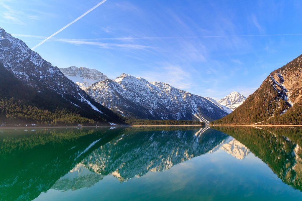A picturesque view of Lake Plansee in Tyrol, Austria with snow-capped mountains reflecting in its crystal-clear waters.