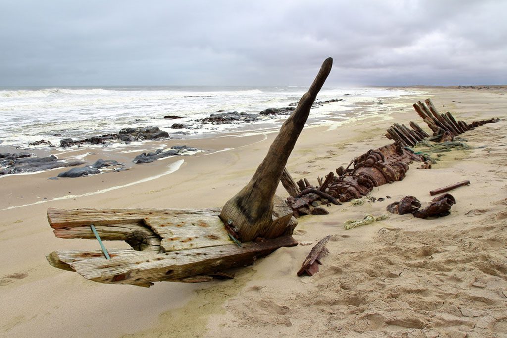 A dramatic photo of a rusty shipwreck stranded on the rocky shores of Skeleton Coast, Namibia, with the vast expanse of the Atlantic Ocean in the background. The ship appears to be partially buried in the sand, with its masts and a few sections of its hull still visible. Photo Contributor: giannimarchetti.