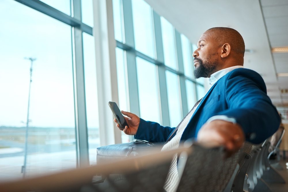 A black man using his phone while waiting at the airport, engaging in travel, business, and networking activities.
