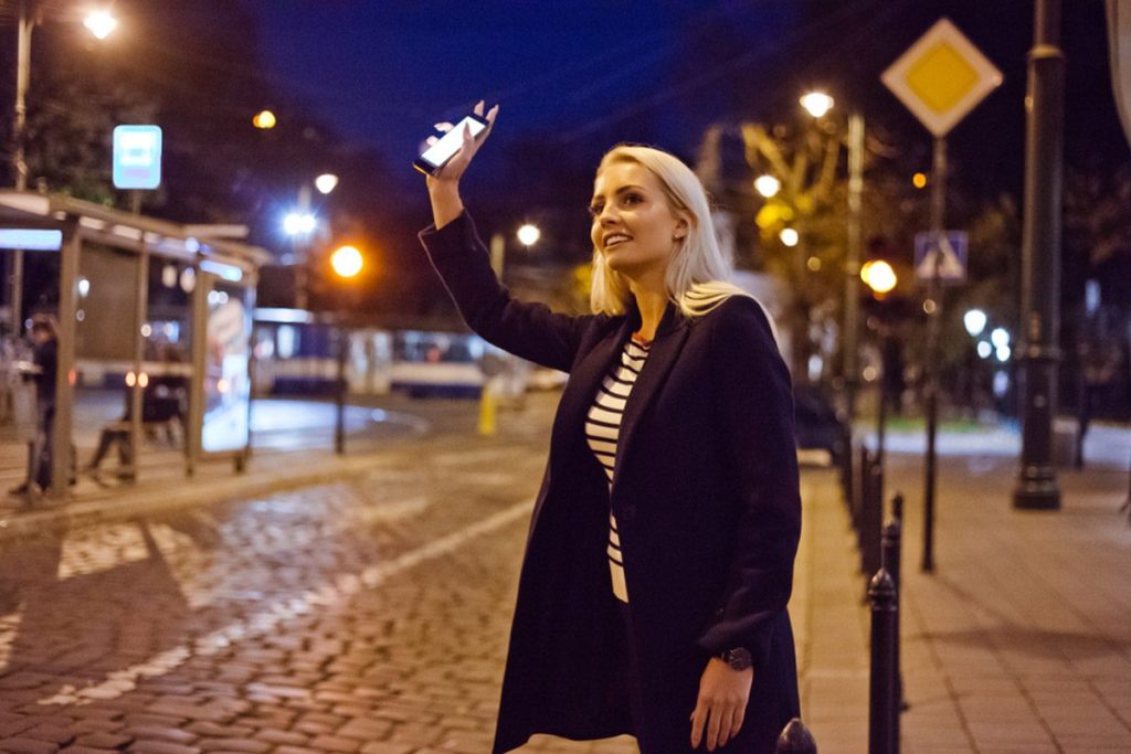Young blonde woman holding a smartphone in the city street, waiting for a taxi or Uber"