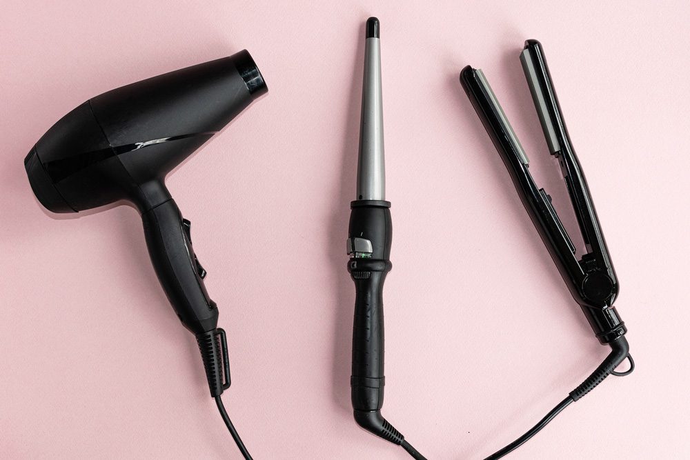 Black hair dryer, curling iron, and hair iron on a pink background.