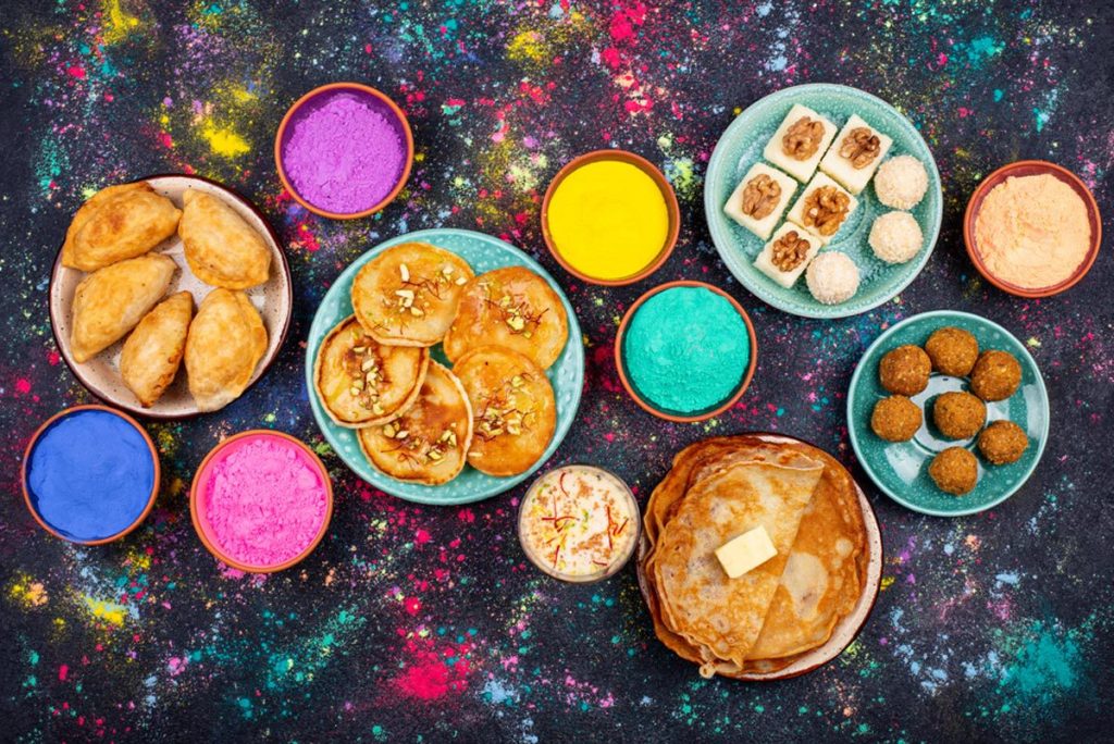 Assortment of mouthwatering Indian sweets and beverages for a Holi party, including laddu, gujiya, puran poli, thandai, malpua, and burfi